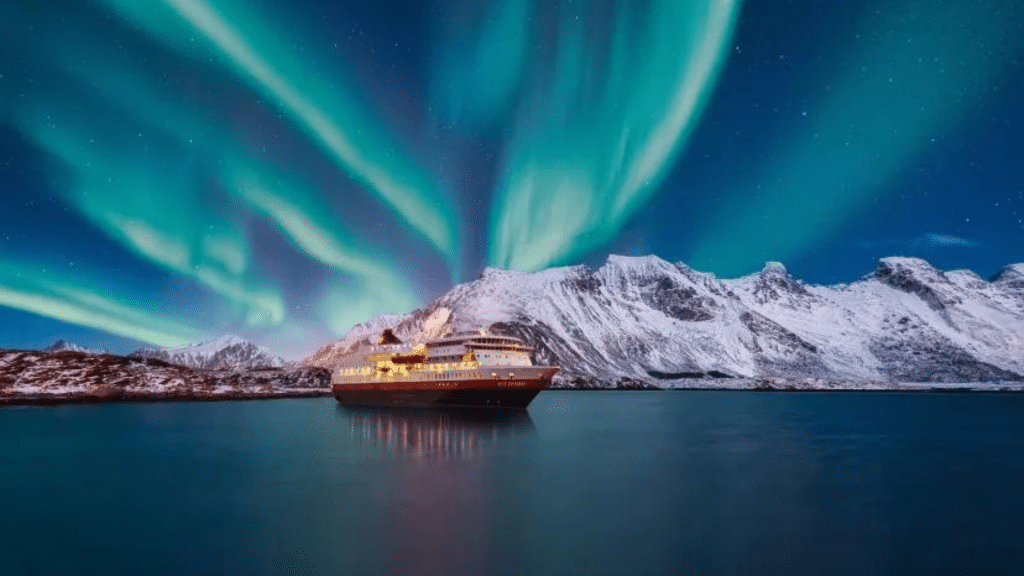 Hurtigruten Schiff unter Nordlichtern vor verschneiter Kueste in Norwegen