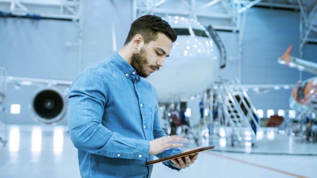 In Big Company Hangar Aircraft Maintenance Engineer Uses Tablet Computer While Standing Near Big New Shiny White Plane.
