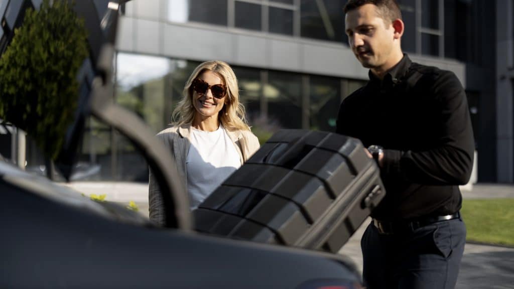 Businesswoman Waits While Chauffeur Packs A Suitcase In Car Trunk