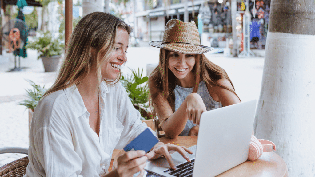 Zwei junge, reisende Frauen laecheln und schauen auf einen Laptop mit Kreditkarte in der Hand